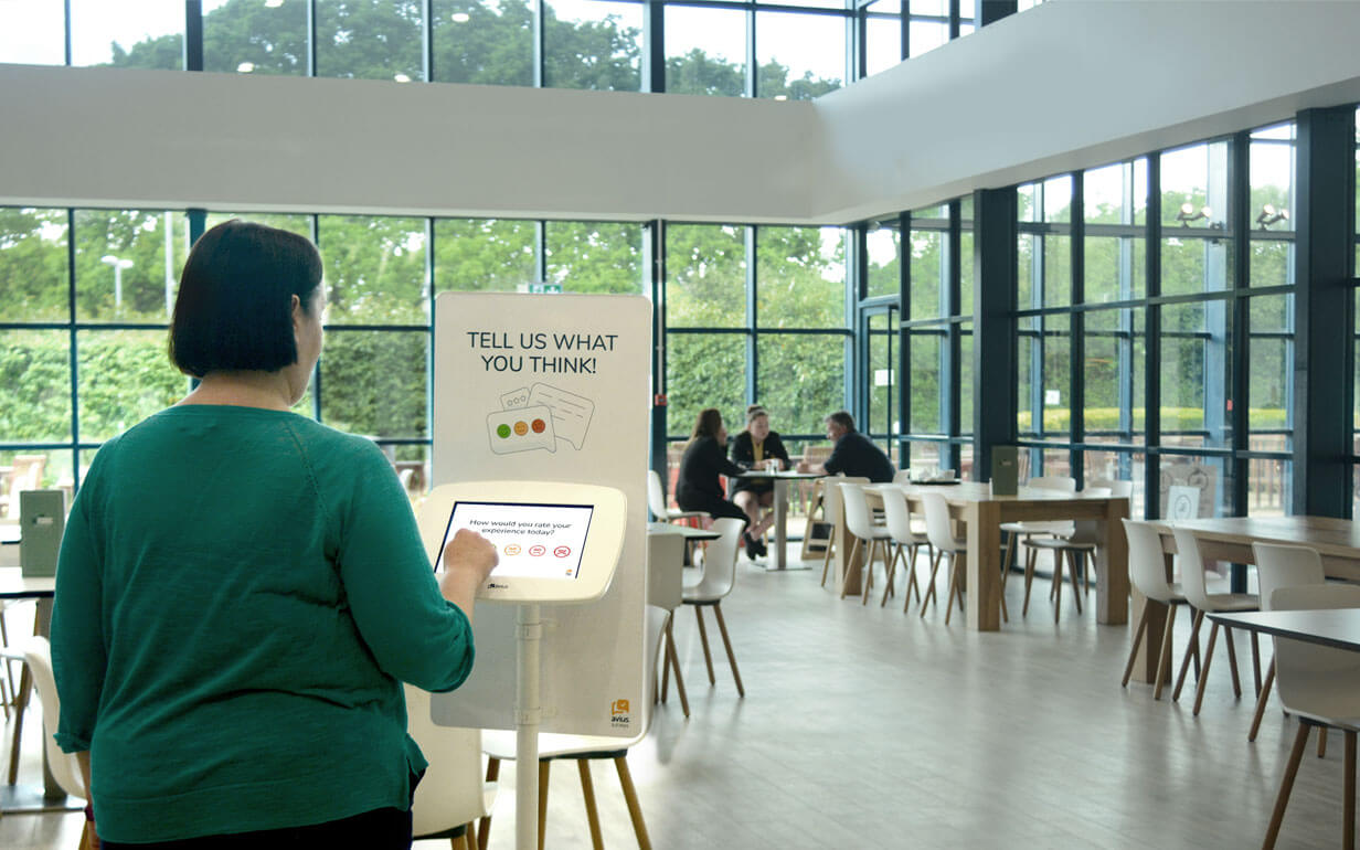 A woman standing in front of a feedback kiosk, tapping on the screen to give feedback regarding a service she has been received. The woman is in a big open space, the coffee of a gym.